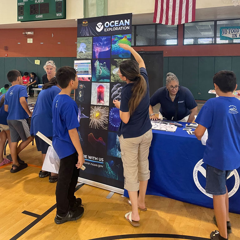 The image shows a gymnasium setting where a small group of people, including children and adults in blue shirts, are gathered around an information booth. The booth is affiliated with NOAA Ocean Exploration, as indicated by the banner and table cover. The banner displays vibrant images of marine life, such as jellyfish and corals, alongside the logo and name of the organization. A woman behind the table is engaged with the visitors, providing information. The scene is lively, with a focus on educational interaction.
