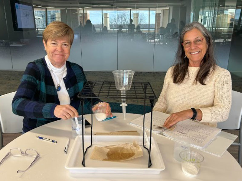 Two women sitting at a table with a filtration setup and various lab materials.