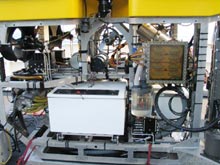 A front view of the ROV Hercules shows the white bio-box, powerful arms, lights, and cameras.