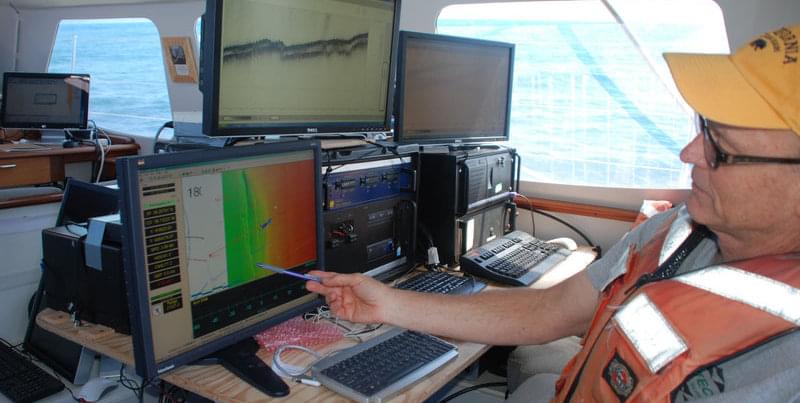 Sam Johnson from USGS points out the location of the San Andreas fault on a map generated by multibeam data to correlate with the incoming seismic data. Bathymetric and backscatter data obtained from multibeam sonar are used to determine seafloor depth as well as indicators of the type of sediment (hard or soft bottom) that is below.