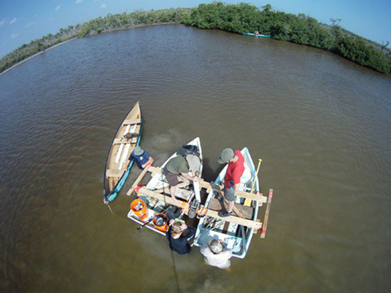 Collecting cores in the field required a team effort and coordination of equipment and people. The team makes use out of the floating coring platform and canoes to collect a core from the West Harbor area.