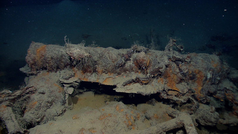 A cluster of large of artifacts near the center of the ship contains three artillery pieces, an anchor and smaller objects. The large gun on top of the pile is a 12 or 18 pounder (meaning it shot a cannon ball weighing 12 or 18 pounds) measuring approximately 10 feet long. Fragments of the gun carriage embedded in the pile suggest this is a deck-mounted center-pivot long gun. Image courtesy NOAA Okeanos Explorer Program.