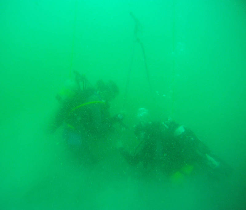 Divers forcing a hydraulic probe into the seafloor. The probe is 10 feet long and is connected by a hose to a water pump on the boat. It jets water out of the end being thrust into the sand.