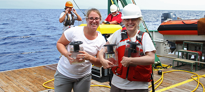Kimberly Puglise (right) and Jana Ash (left) carry sample bins full of specimens from the remotely operated vehicle on deck into the lab for processing.