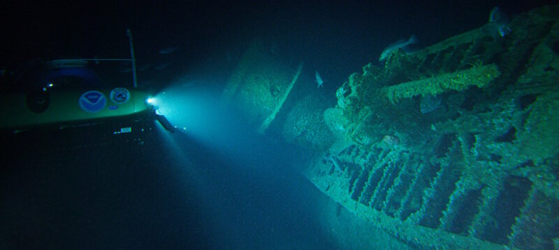 A submersible shines its lights on the wreck of U-576 lying on its starboard side, showing the submarine's conning tower and the deck gun in the foreground.