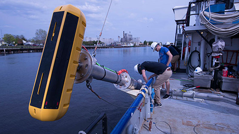 University of Delaware sonar technicians Kenny Haulsee and Peter Barron route cables from the echosounder (yellow device in foreground) along a pole mount used to lower and secure the echosounder along the side of the vessel. The team installed and tested the entire sonar system while dockside to ensure proper operation before getting underway within the survey area.