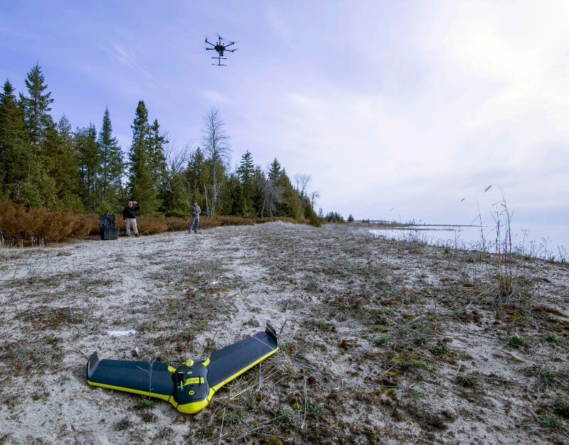 A fixed-wing senseFLY eBee RTK aircraft rests on the ground as a DJI Matrice 600 takes off in the background. NOAA, operating the eBee system, works with partners Oceans Unamanned and Trumball Unmanned to survey shallow-water portions of the Thunder Bay National Marine Sanctuary.