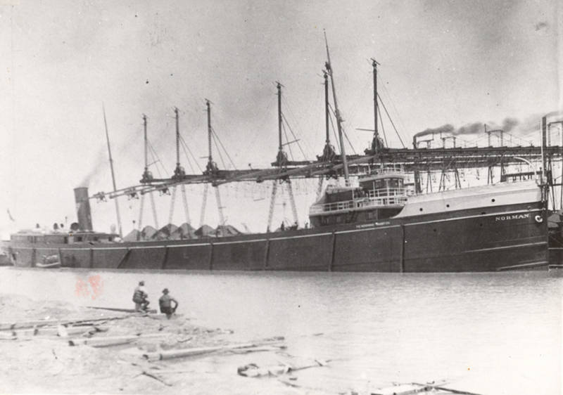 Historical image of Norman at a loading dock, profile of the starboard side of the freighter.