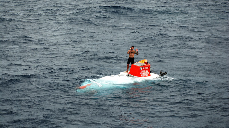 Swimmer and R/V Atlantis crew member Ronnie Whims stands atop Alvin after it surfaced for recovery. Ronnie communicates with the team still inside the sphere via phone until the sub is secured and ready to be hauled aboard.
