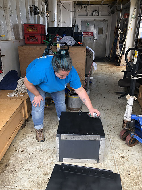 Alexis Weinnig spray paints the top of the bioboxes that will be used to safely store live coral samples collected during dives. The black matte paint helps to reduce glare that could interfere with the video cameras mounted above the sample basket.