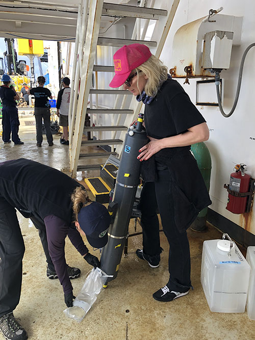 Kate Segarra holds a 12-liter Niskin bottle that had been sent down on ROV Jason while Cheryl Morrison (USGS) collects the water from it. Cheryl will then analyze the water for the presence of environmental DNA.