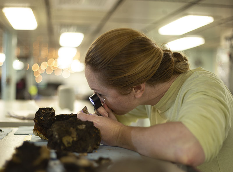 Amy Gartman, U.S. Geological Survey Research Oceanographer and chief scientist for the Escanaba Trough expedition, examines a mineral sample with a hand lens aboard Research Vessel Thomas G. Thompson during the Escanaba Trough: Exploring the Seafloor and Oceanic Footprints expedition. Image courtesy of U.S. Geological Survey.