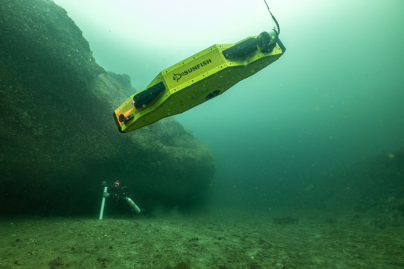 Cave diver Tamara Adame collects sediment samples beneath an overhang in southeast Alaska during 2023 field work for the Our Submerged Past project while autonomous underwater vehicle SUNFISH documents the sampling process and creates a 3D map of the sample site.