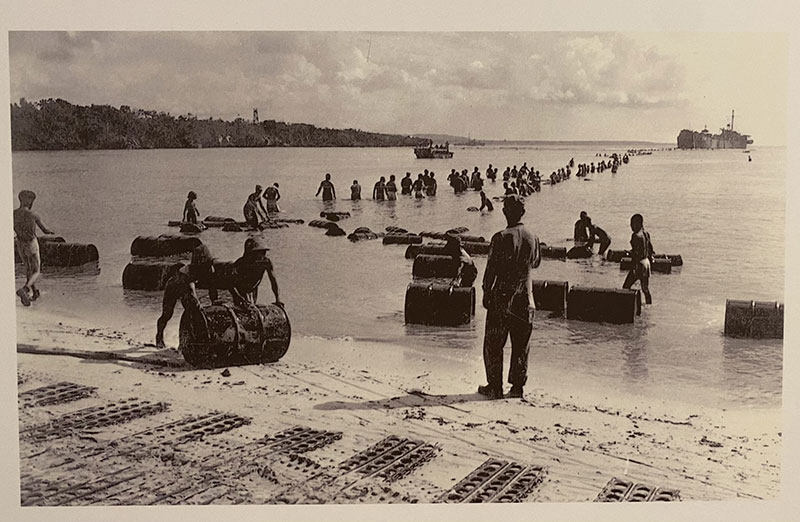 U.S. soldiers unloading supplies on a Pacific Island beach during World War II with pierced steel planking (PSP) in the sand in the foreground.