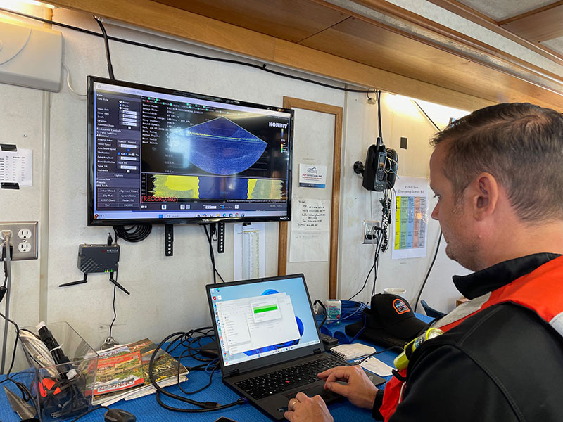 A man in a life jacket operates a laptop in front of a wall-mounted monitor displaying radar data aboard a ship.