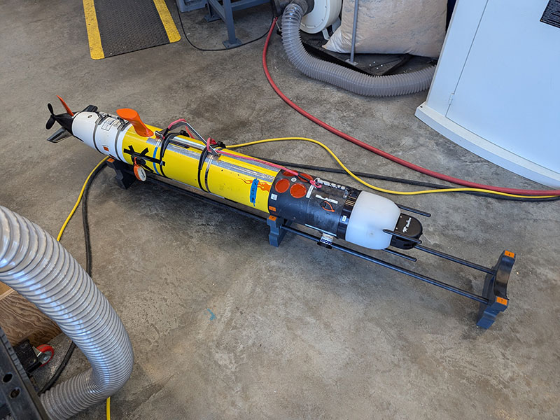 Autonomous underwater vehicle with yellow, black, and white sections on a concrete floor surrounded by hoses and cables.