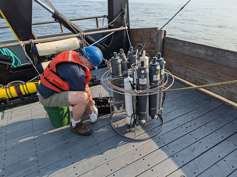 A man in safety gear works attentively with scientific equipment on a ship’s deck near the ocean.