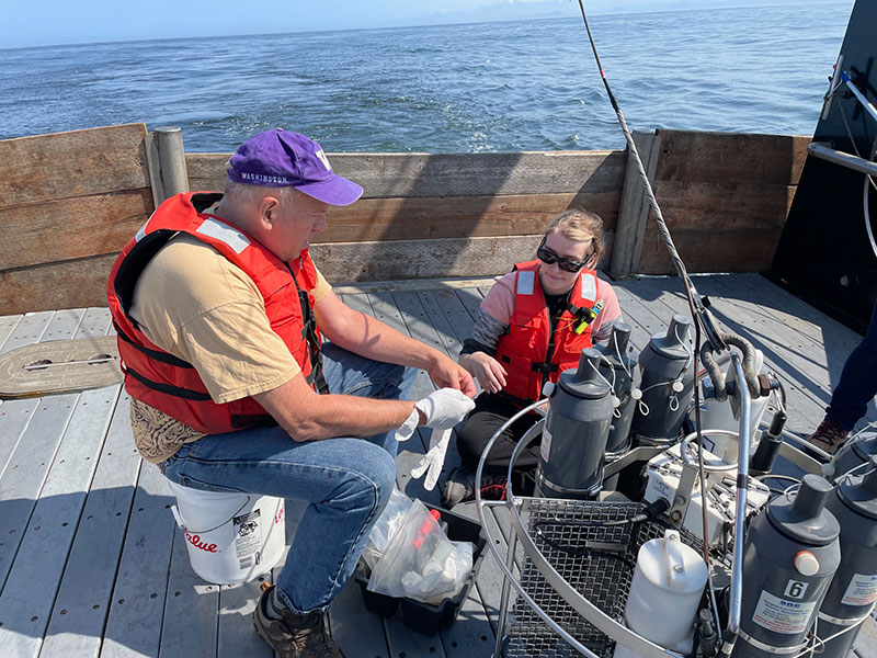 Two people working on a boat deck with scientific equipment and wearing life vests.