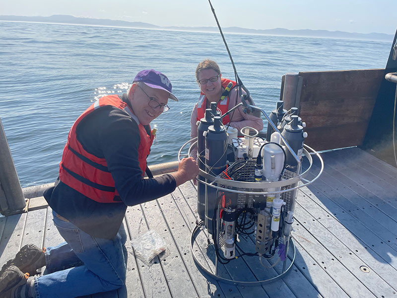Two people on a boat deck working with oceanographic equipment, wearing life vests, with an ocean backdrop.