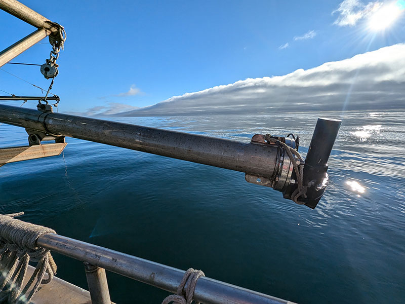A metal pole extends over calm ocean waters, with clouds and sunlight above.