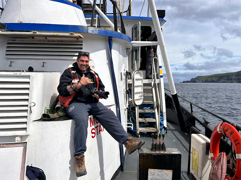 The image depicts a smiling man sitting on the edge of a section of a boat. He is giving a thumbs-up gesture while holding a camera. The man is wearing a black jacket, a red life vest, gray sweatpants, and brown work boots. The weather appears partly cloudy. The boat has a predominantly white structure with blue trim. Behind the man, there is a set of white stairs leading to another level of the boat. The open water is visible on the right side of the image, with a coastline featuring green hills in the distance. A red life preserver ring is mounted on the railing at the edge of the boat.