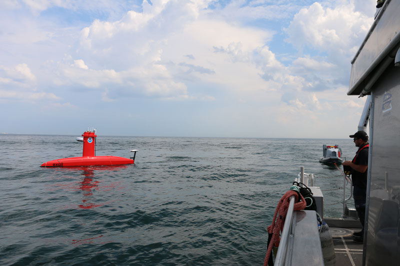 A red unmanned surface vessel on the ocean with a person controlling it from a nearby ship.