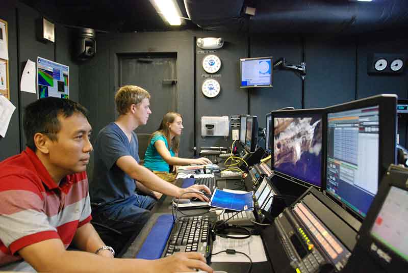 Typical night time mapping watch in the control room. Back to front: Senior Survey Technician Elaine Stuart monitors incoming data and adjusts settings as necessary; ROV team member, Tom Kok, does double duty by processing mapping data as it is collected; Major Dian Adrianto provides hourly status updates about ship operations to shore via intercom and the Eventlog.