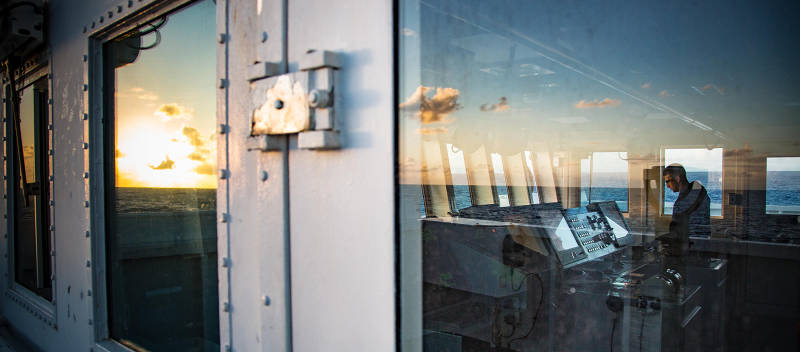 An ocean sunset is seen in the reflection of the port windows on the Bridge of NOAA Ship Okeanos Explorer. Inside the Bridge, ENS Chris Licitra examines the shipʻs radar for contacts.