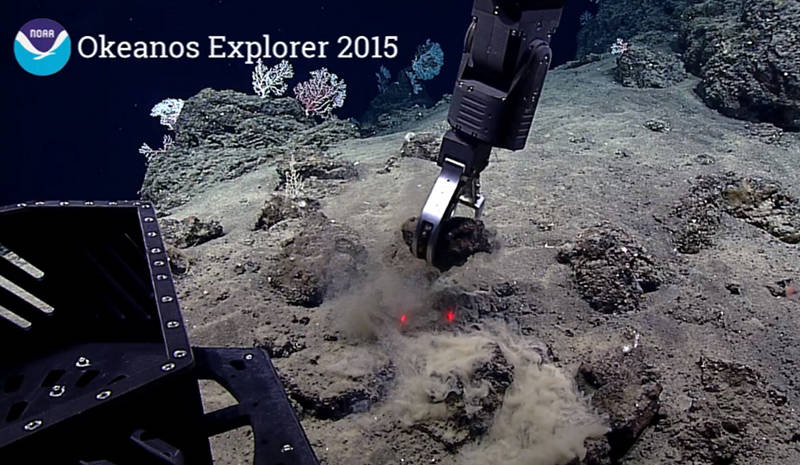 ROV arm collecting a rock sample from the 1868 lava flow in order to confirm the age of the substrate using geochemical ratio and weathering analysis. The laser points (red dots) are 10 centimeters (~4 inches) apart and are used by scientists to determine the sizes of things that they are seeing.