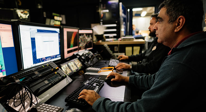 Expedition Mapping Lead, Mashkoor Malik, works on planning the mapping lines the ship will run today since the weather is too poor to dive. Behind him, Survey Technician Charlie Wilkins edits recently acquired multibeam data.