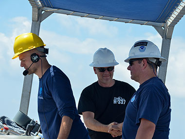 NOAA Ship Okeanos Explorer Operations Officer, LT Aaron Colohon, shakes Commanding Officer, CDR Eric Johnson’s hand following successful recovery of the remotely operated vehicle (ROV). All power and communications to the vehicle were lost during the dive, making recovery more complicated than usual. Next to them, Global Foundation for Ocean Exploration ROV Dive Supervisor, Dan Rogers, oversees operations on the aft deck following recovery.