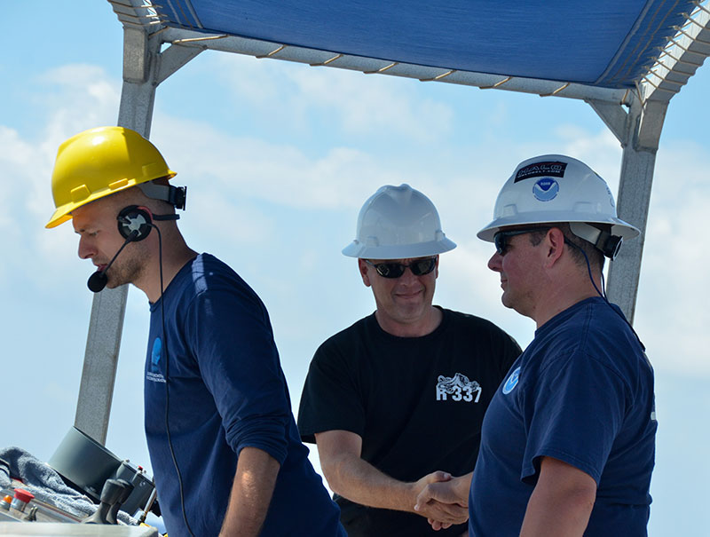 NOAA Ship Okeanos Explorer Operations Officer, LT Aaron Colohon, shakes Commanding Officer, CDR Eric Johnson’s hand following successful recovery of the remotely operated vehicle (ROV). All power and communications to the vehicle were lost during the dive, making recovery more complicated than usual. Next to them, Global Foundation for Ocean Exploration ROV Dive Supervisor, Dan Rogers, oversees operations on the aft deck following recovery.