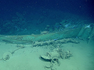 First view of the wreck’s bow outlined by the remnant copper sheathing, with one hawse pipe in foreground and another just aft of the stempost at right. Also visible are white colonies of the stony coral Lophelia pertusa (on the stempost and at far left) and numerous duckbill eels.