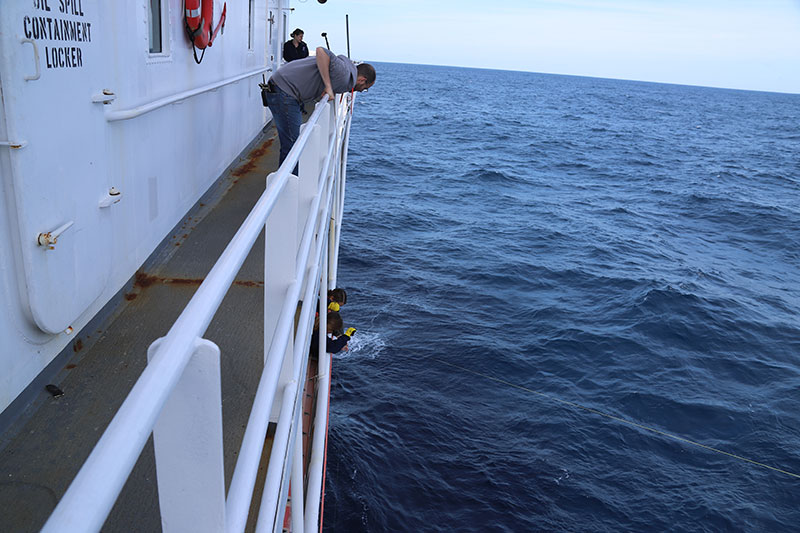 Image of the calibration team attaching the calibration sphere to the three spectra lines on the starboard side of NOAA Ship Okeanos Explorer. The yellow spectra line from the starboard aft downrigger can be seen in the right portion of the image.