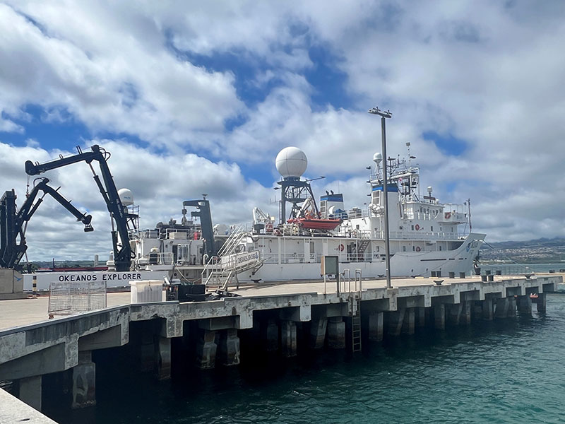 Research vessel 'Okeanos Explorer' docked at a pier with cranes and radar equipment on deck under a partly cloudy sky.
