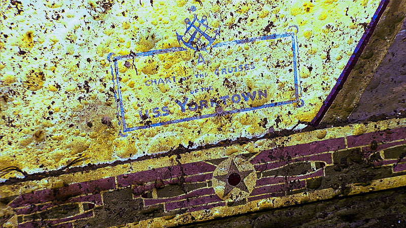 Yellowish corroded metal surface with blue stamped text and maroon stripes on the U.S.S. Yorktown's hull section.