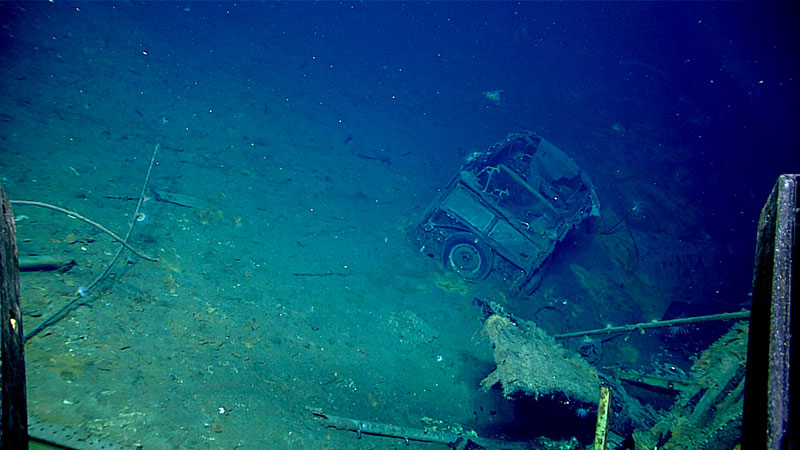 A sunken vehicle partially buried on the ocean floor with surrounding debris.