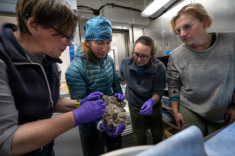 Expedition biology science lead Rhian Waller, geology science lead Jen Aschoff, sample data manager Anna Lienesh, and data manager Jennifer Green examine a rock sample collected during Dive 04 of the Seascape Alaska 3 expedition.